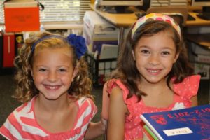 Two smiling young girls sitting together, holding books.