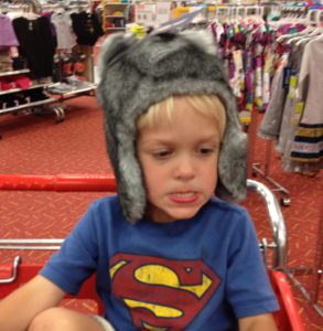 Young boy wearing a gray furry hat and Superman shirt sitting in a shopping cart.