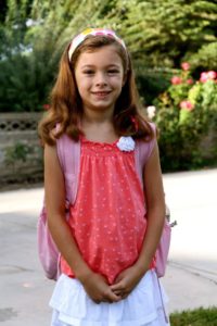 A smiling girl in a pink dress stands outdoors with flowers in the background.