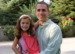 A man and young girl smiling outdoors with greenery behind them.