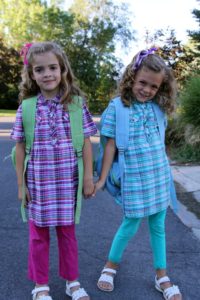 Two young girls holding hands, smiling outdoors in colorful plaid dresses.