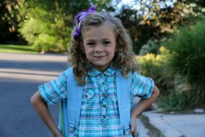 A smiling young girl with curly hair and a purple bow outdoors.