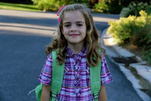 Smiling girl with curly hair wearing a pink plaid shirt and green backpack outdoors.